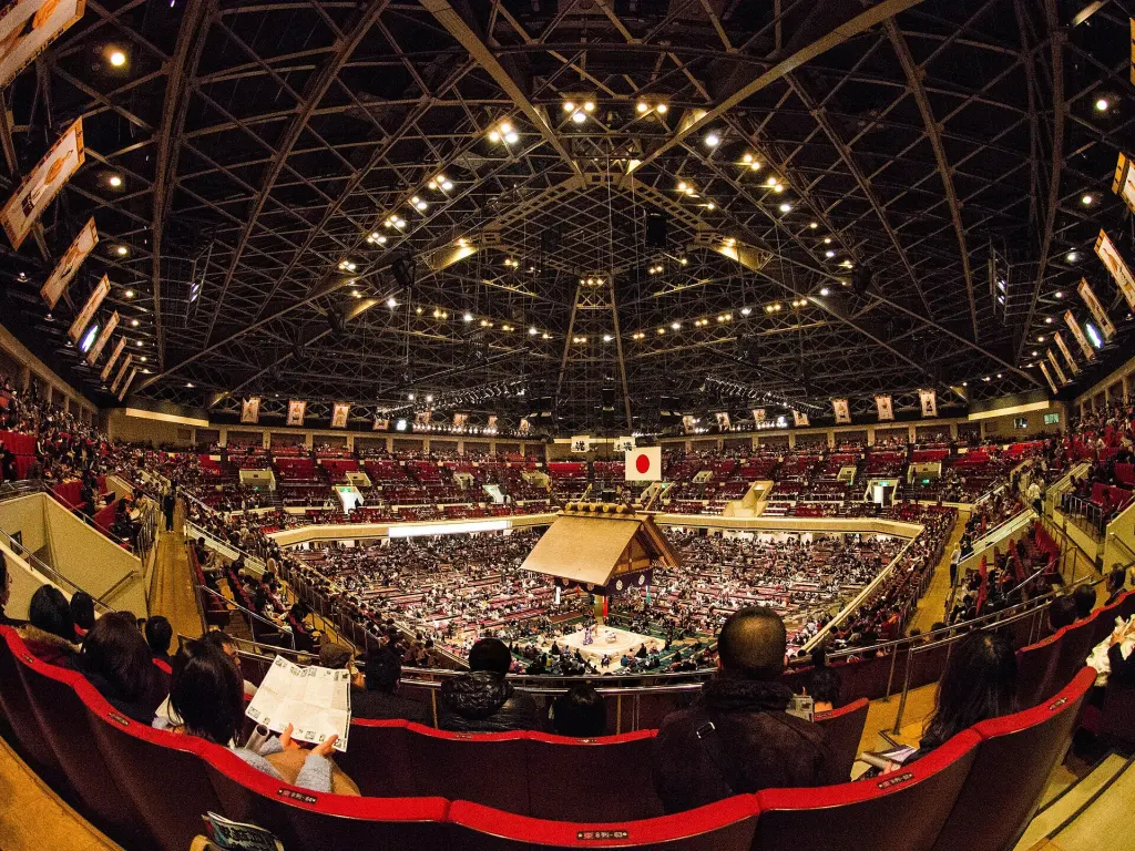 Crowded Ryōgoku Kokugikan with a raised sumo ring beneath the suspended roof