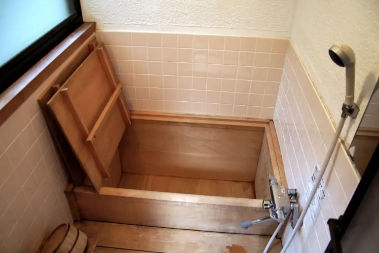 Deep wooden Japanese bathtub with a lid and hand shower in a tiled bathing room