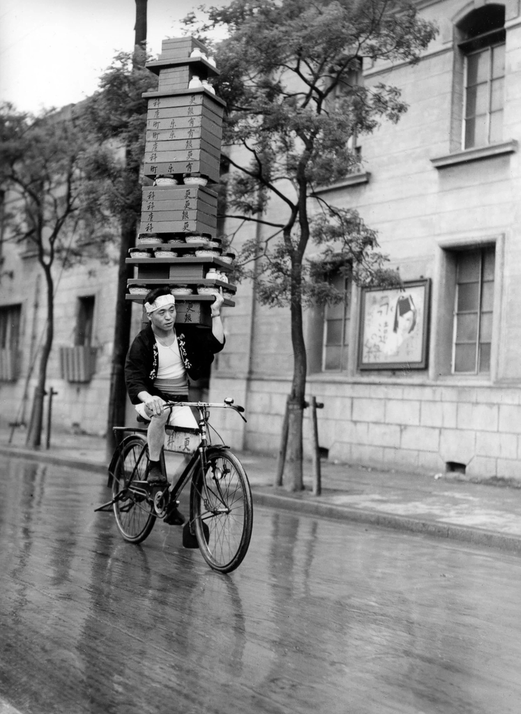 Black-and-white photograph of a bicycle courier carrying a tall stack of soba bowls in Tokyo