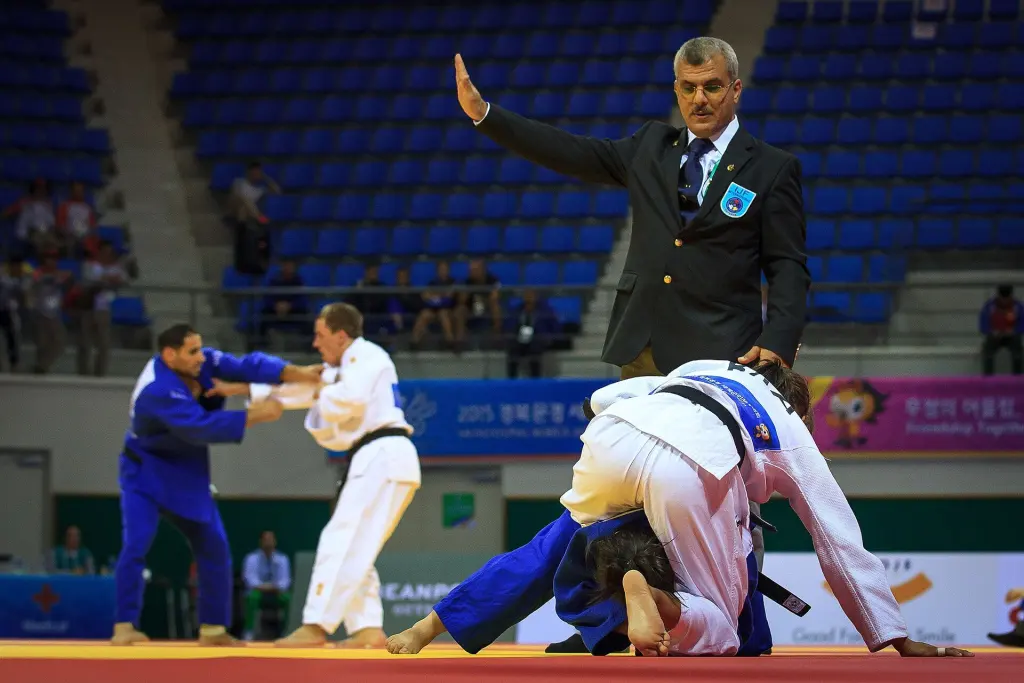 A Judo referee signals while two judoka continue ground action